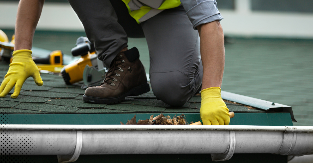 Worker wearing brown boots and yellow gloves cleaning a rooftop gutter filled with debris and leaves.