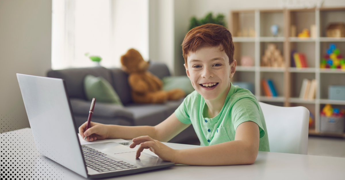 Smiling boy using a laptop at home with a notebook and pencil.